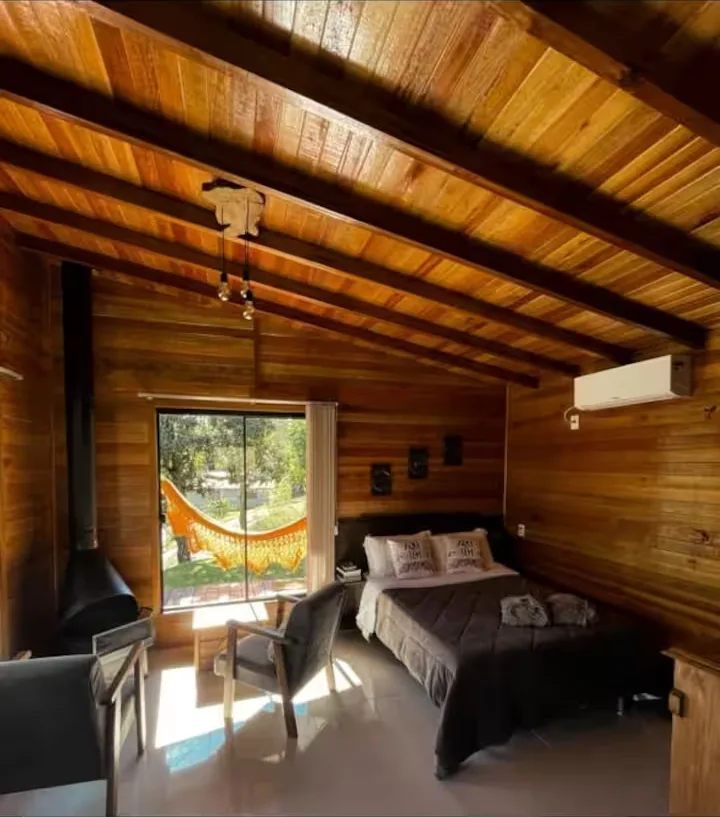 Quarto do chalé com cama de casal, lareira e rede na varanda vista pela janela